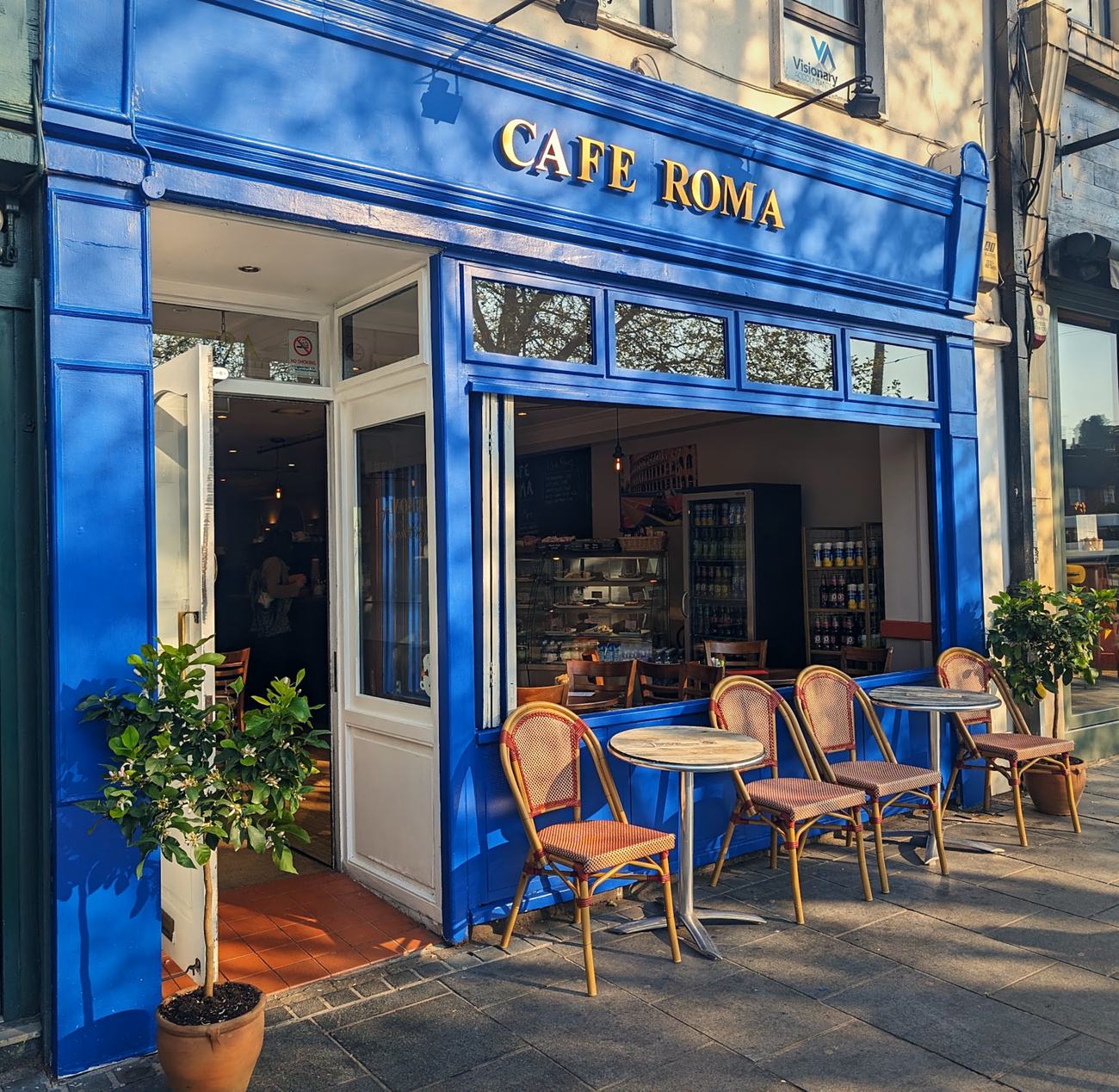 The blue shopfront of Cafe Roma on St Peter's Street, with gold lettering, pavement tables and lemon trees